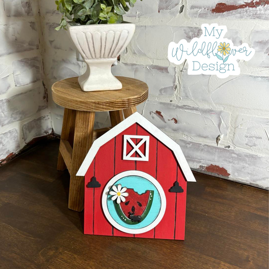Red barn-shaped decorative item with a white roof on a wooden surface, next to a plant on a small wooden stool.